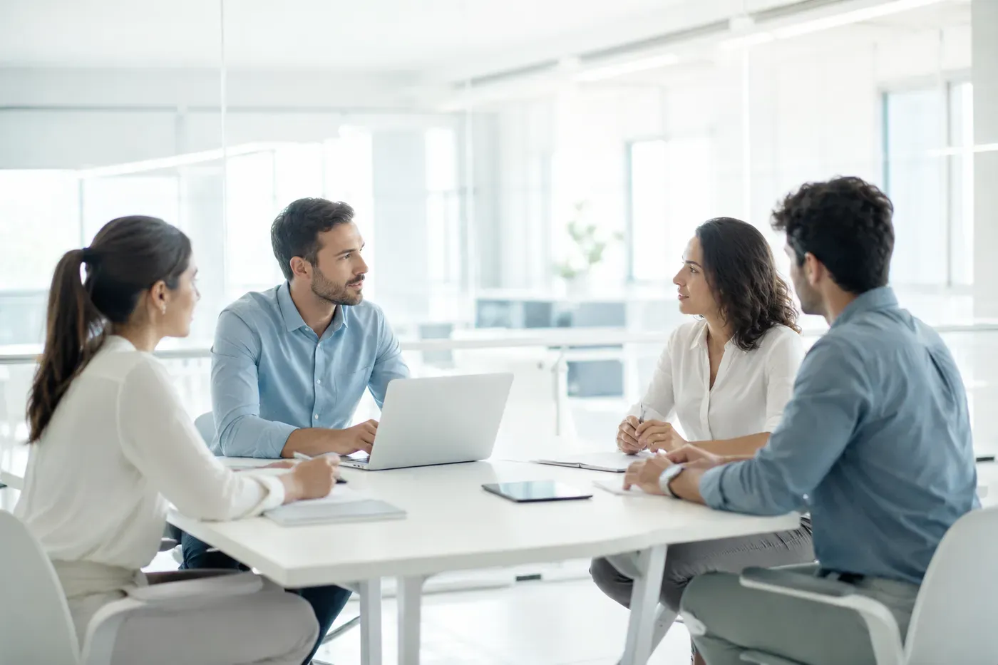 Four colleagues at a table in a bright office with laptops and tablets, talking to each other