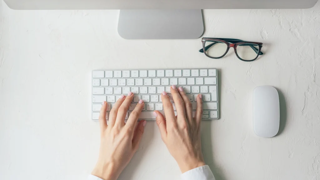Manos femeninas tecleando en un teclado blanco junto a unas gafas y un ratón